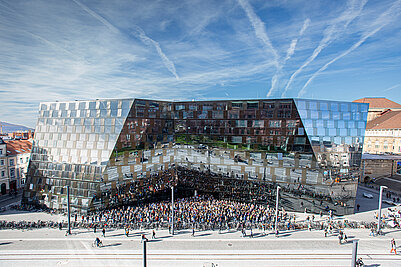 Frontalansicht des neuen Gebäudes der Universitätsbibliothek Freiburg. An einem sonnigen Tag mit blauem Himmel spiegelt sich die Umgebung in der Fassade wider. Viele Menschen stehen vor dem Gebäude und halten eine Schweigeminute für die Opfer der Terroranschläge in Paris im November 2015.